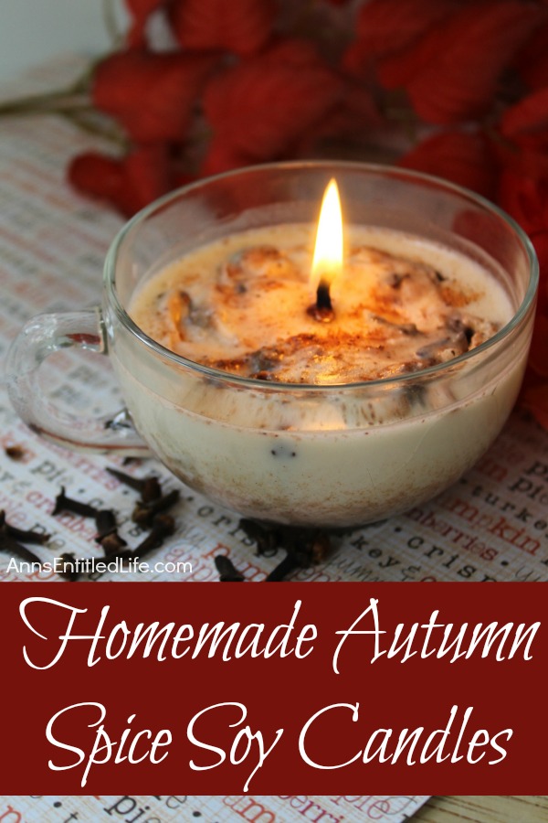 An overhead view of a lit homemade autumn spice candle in a clear teacup on a words sheet. There is red floral in the background.