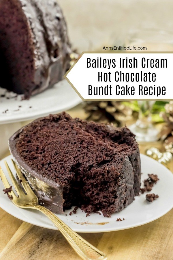 A slice of Baileys Irish Cream Hot Chocolate Bundt Cake laying on its side on a white plate, a bite is missing, There is a gold fork on the left side of the plate. The remaining bundt cake is in the upper left. These are set upon a beige placemat.