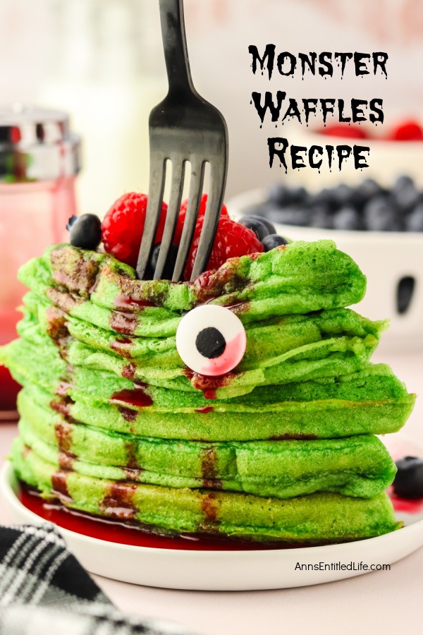 A stack of green waffles drenched in red syrup on a white plate. The waffles are topped with berries and candy eyes, and a fork is descending into the pile.