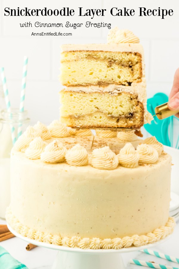 A piece of Snickerdoodle cake is being lifted on a gold cake lifter from the whole Snickerdoodle cake, set on a white cake stand, below.