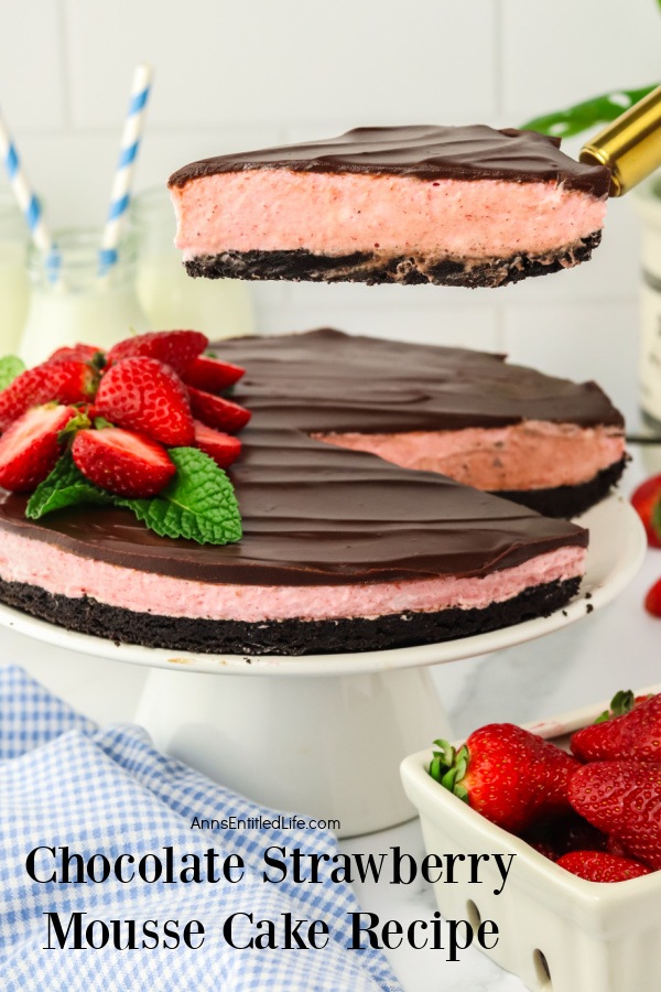 A slice of Homemade Chocolate Strawberry Mousse Cake is being lifted from the whole cake on the white cake stand. There is a pint of fresh strawberries in the lower right.